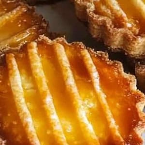 May include: Close-up of several golden-brown tarts with a fluted crust and a glossy, glazed topping. The tarts have a ribbed pattern on top, suggesting a fruit filling. The baked goods are arranged closely together.