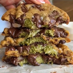 以下が含まれることがあります： 緑色のフィリングと溶けたチョコレートが入った、チョコレートチップクッキーの山。クッキーは、チョコレートチップと塩がまぶされた、金色がかった茶色です。緑色のフィリングはクッキーの層の間に見え、溶けたチョコレートが滴り落ちています。
