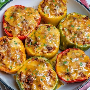 May include: A top-down view of a white pan filled with seven stuffed bell peppers. The peppers are red, yellow, and green, filled with a meat mixture and topped with melted cheese and parsley.