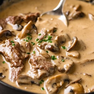 Pode incluir: Close-up de carne stroganoff numa panela. Pedaços tenros de carne e cogumelos fatiados estão imersos num molho cremoso castanho claro. Cebolinho fresco polvilhado por cima. Uma colher de prata está no prato.