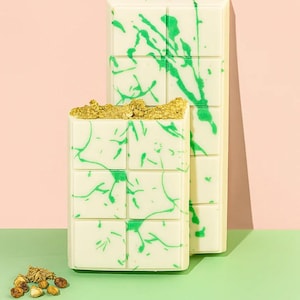 Könnte beinhalten: Zwei weiße Schokoriegel mit grünen Spritzmustern. Ein Riegel ist mit einer Ansammlung goldbrauner Stücke belegt. Die Riegel befinden sich auf einem grünen und rosa Hintergrund. Ein kleiner Haufen goldbrauner Stücke befindet sich im Vordergrund.