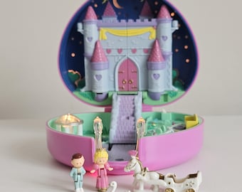 Puede incluir: Un castillo de juguete en forma de corazón de color morado que se abre para revelar un interior iluminado con una escalera central, torres y un fondo de cielo nocturno. Incluye pequeñas figuras de una persona, un personaje real, un cisne y un carruaje.