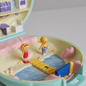Op de afbeelding: Een lichtblauwe, schelpvormige speelset met een miniatuur strandscène en een gele houten brug over blauw water. Twee kleine plastic figuurtjes, één met blond haar in een rode outfit en één met een gele hoed en geel shirt, staan op het zand.