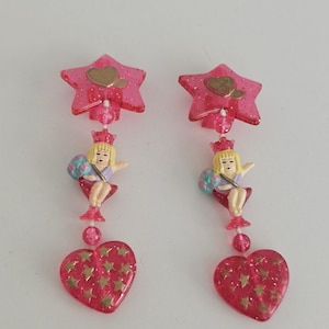 May include: A pair of pink dangle earrings. Each earring features a pink star-shaped top with a gold heart, a miniature princess figure, and a heart-shaped bottom with gold stars. The earrings are made of plastic.