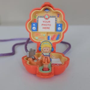 May include: A vintage, orange, flower-shaped toy with a miniature doll inside. The toy opens to reveal a room with a bed, a TV, and a DJ booth. The top section has a stage with the text "YOUR PHOTO HERE". A purple cord is attached.