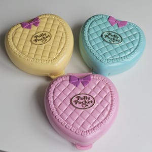 Op de afbeelding: Drie hartvormige Polly Pocket compactdoosjes in geel, lichtblauw en roze. Elk plastic doosje heeft een gewatteerde textuur, een paarse strik aan de bovenkant en een centraal ovaal plaatje met de tekst Polly Pocket en een hartje.