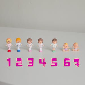 May include: A collection of seven small plastic figurines arranged in a row, labeled with the numbers 1 through 7 in pink. The figures include five standing children in patterned outfits and two seated infants wearing diapers, all with painted facial features.