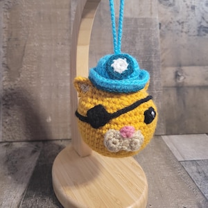 May include: A crocheted yellow cat with a black eye patch and a blue hat with a white emblem. The cat has a pink nose and a beige mouth. It is hanging from a wooden stand with a blue string.