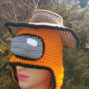 May include: An orange crochet hat with a gray visor and a white and gray face design. The hat has a brown brim and black trim. The hat is on a mannequin head.