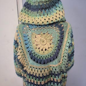 Könnte beinhalten: Handgefertigte Kapuzenjacke aus Häkelarbeit im Granny-Square-Muster. Die Jacke hat eine Farbpalette aus Blau-, Grün- und Beigetönen. Kapuze und Körper sind in einem kreisförmigen Design gestaltet und schaffen ein einzigartiges und gemütliches Kleidungsstück.
