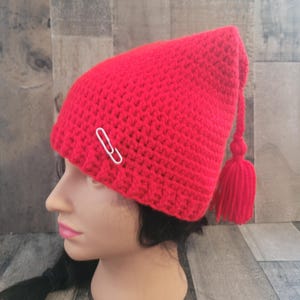 May include: A bright red crocheted hat with a pointed top and a tassel. A silver paperclip is attached to the hat. The hat is displayed on a mannequin head.