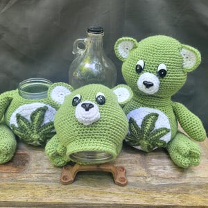 May include: Handmade green crochet bear with white accents and a cannabis leaf design on the belly. The bear is sitting next to a jar and a glass bottle. The bear is made of soft yarn and has black eyes.