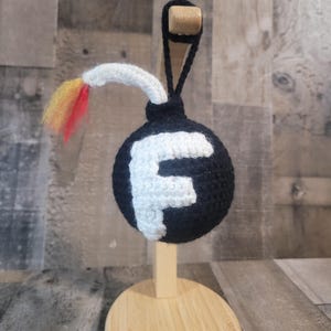 May include: A black crocheted bomb ornament with a white "F" on the front and a white fuse with red, yellow, and orange accents. The ornament is displayed on a wooden stand.