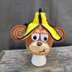 May include: A handmade crochet monkey hat with a banana peel on top. The hat is brown, tan, yellow, and black, with large white eyes and a smiling face. The hat is displayed on a white stand.