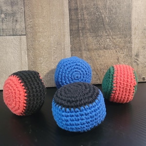 May include: Four colorful crochet juggling balls. One ball is red and black, another is blue, a third is blue and black, and the last is green and red. The balls are arranged on a dark surface with a wooden background.
