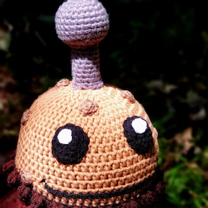 May include: A handmade crochet hat in the shape of a whimsical creature. The hat features a tan body with brown accents, large black eyes with white highlights, and a purple antenna. The bottom edge is trimmed with a dark brown scalloped design.