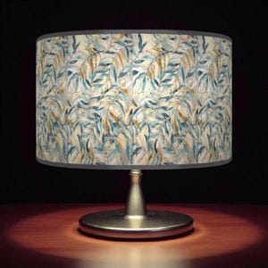 May include: A table lamp with a cylindrical lampshade patterned with blue and gold leaves on a cream background. The lampshade has a grey border. The lamp has a silver-coloured base and stem.