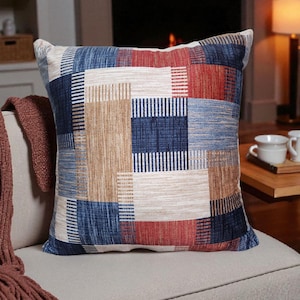 May include: Square cushion with a patchwork design in shades of blue, red, and tan. The cushion is placed on a neutral-coloured sofa. The cushion's pattern features rectangular blocks with various textures and patterns.