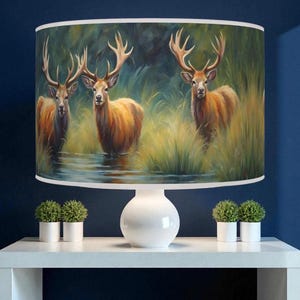 May include: A white table lamp with a cylindrical lampshade depicting three deer in a marsh. The deer have large antlers and are surrounded by tall grass. The background is a dark, moody landscape.
