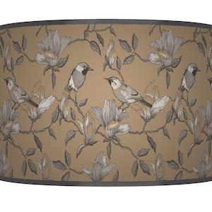 May include: A cylindrical lampshade with a tan background, featuring a repeating pattern of grey birds perched on flowering branches. The flowers are grey and white, and the rim is a dark grey.