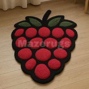May include: A raspberry-shaped rug with a black border, red berry-like circles, and green leaf accents. The rug has the text "Mazerugs" in white. The rug is placed on a light-colored wooden floor.