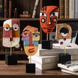 Peut inclure: Quatre sculptures de visages abstraits au design moderne. Chaque sculpture est montée sur une tige dorée et une base carrée noire. Les visages présentent une variété de couleurs, dont le rouge, l'orange, le jaune et le bleu.