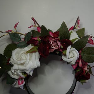 Può includere: Una corona floreale con rose bianche e bordeaux, foglie verdi e accenti rosa. Le rose sono adornate con piccoli abbellimenti scintillanti. La corona ha una base nera e dettagli color oro.