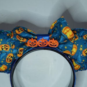 Puede incluir: Una diadema con dos orejas circulares y un lazo central, cubiertos de tela azul verdoso con un estampado de calabazas naranjas, sombreros de bruja azules y estrellas amarillas. El lazo está decorado con tres pequeños dijes de calabaza.