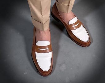 May include: Brown and white leather penny loafers. The shoes have a brown leather upper with a white vamp and a brown leather sole. The shoes are worn with tan pants.