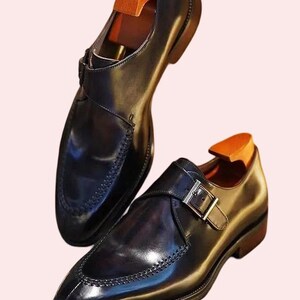 Könnte beinhalten: Ein Paar schwarze Lederschuhe mit Monk-Strap-Verschluss und poliertem Finish. Die Schuhe haben eine dekorative, abgesteppte Zehenkappe und einen silberfarbenen Schnallenverschluss. Schuhspanner aus Holz sind eingesetzt.