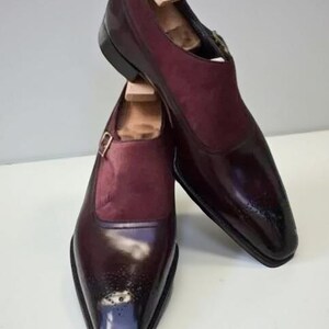 May include: A pair of burgundy leather dress shoes with a monk strap buckle closure. The shoes have a glossy finish on the toe and a suede panel on the upper. Wooden shoe trees are inserted inside.