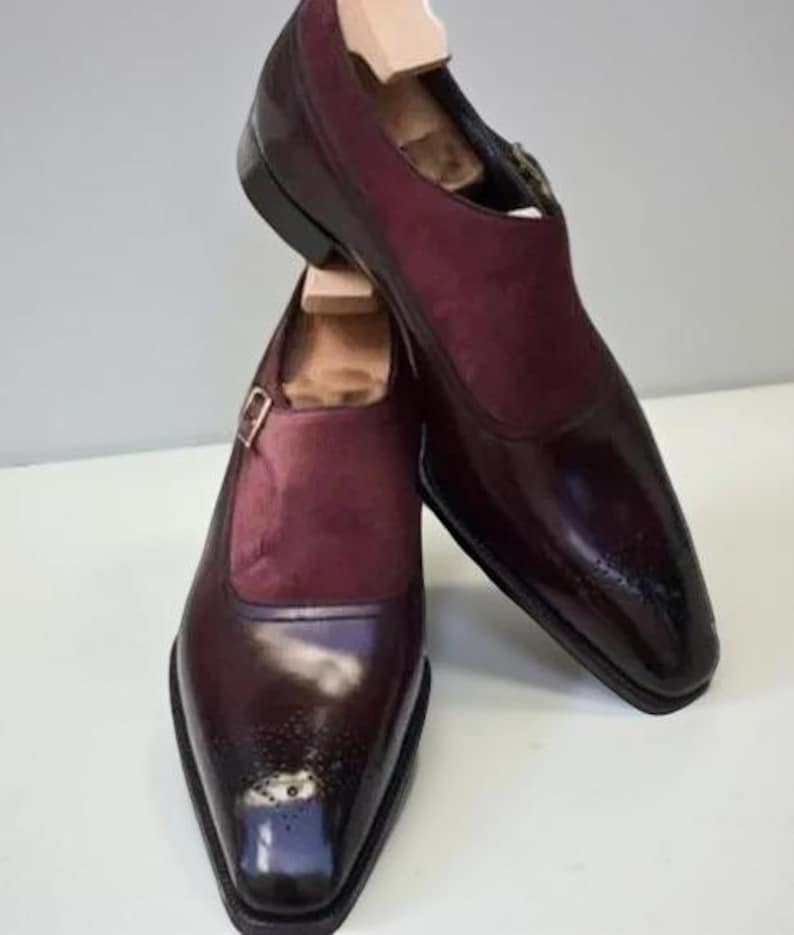 May include: A pair of burgundy leather dress shoes with a monk strap buckle closure. The shoes have a glossy finish on the toe and a suede panel on the upper. Wooden shoe trees are inserted inside.