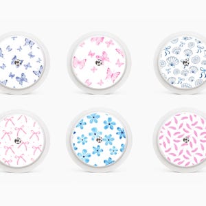 May include: Six white, circular objects with decorative patterns. Designs include blue butterflies, pink butterflies, seashells, pink bows, blue flowers, and pink feathers. Each design has a small black ladybird accent.