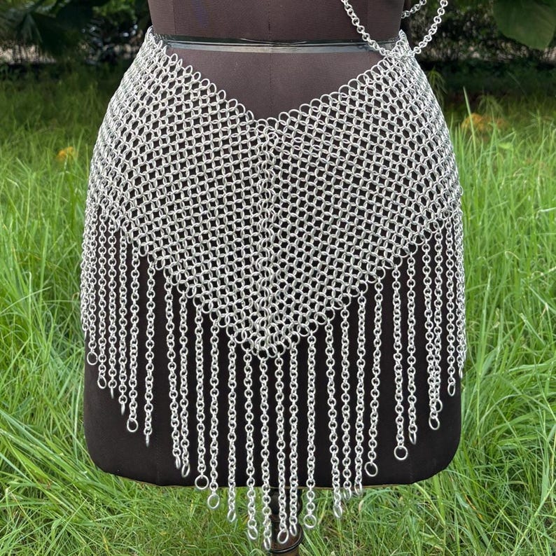 Puede incluir: Una falda de cota de malla plateada con un patr&oacute;n geom&eacute;trico y flecos de cadena colgantes. La falda se exhibe en un maniqu&iacute; negro, sobre un fondo de hierba verde. La cota de malla est&aacute; hecha de peque&ntilde;os anillos de metal entrelazados.
