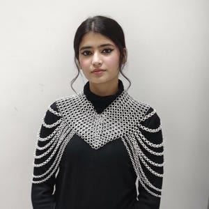 Puede incluir: Una pieza de hombro de cota de malla plateada con un diseño geométrico. La cota de malla está drapeada sobre una camiseta negra de manga larga. La pieza del hombro tiene un cuello alto y cadenas en cascada.