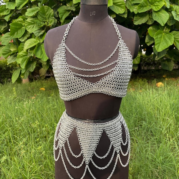 May include: A silver chainmail bikini set displayed on a mannequin. The set includes a halter top with multiple chain strands and a skirt with a decorative chain fringe. The mannequin is set against a backdrop of green foliage.
