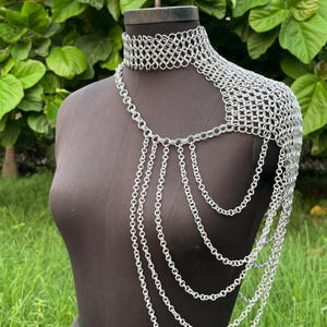 Puede incluir: Una pieza de hombro de cota de malla plateada con un cuello tipo gargantilla y cadenas en cascada. El intrincado dise&ntilde;o se muestra en un maniqu&iacute; sobre un fondo de follaje verde, mostrando la artesan&iacute;a de la joyer&iacute;a.