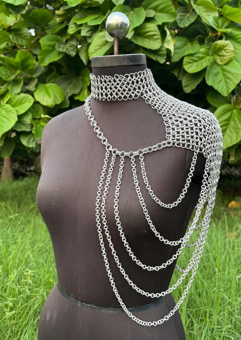 Puede incluir: Una pieza de hombro de cota de malla plateada con un cuello tipo gargantilla y cadenas en cascada. El intrincado dise&ntilde;o se muestra en un maniqu&iacute; sobre un fondo de follaje verde, mostrando la artesan&iacute;a de la joyer&iacute;a.