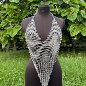 Puede incluir: Un top halter de cota de malla plateada con diseño de cuello en V. El top se exhibe en un maniquí negro sobre un fondo de follaje verde y hierba. La cota de malla está hecha de pequeños anillos de metal entrelazados.