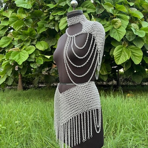 Puede incluir: Un conjunto de cota de malla plateada exhibido en un maniqu&iacute;. El conjunto incluye una pieza para el hombro con cadenas en cascada, un collar&iacute;n y una falda con dobladillo con flecos. El maniqu&iacute; se encuentra sobre un fondo de follaje y hierba verdes.