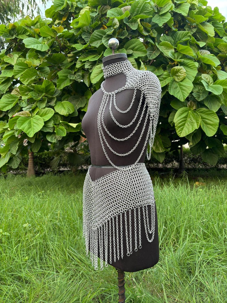 Puede incluir: Un conjunto de cota de malla plateada exhibido en un maniqu&iacute;. El conjunto incluye una pieza para el hombro con cadenas en cascada, un collar&iacute;n y una falda con dobladillo con flecos. El maniqu&iacute; se encuentra sobre un fondo de follaje y hierba verdes.