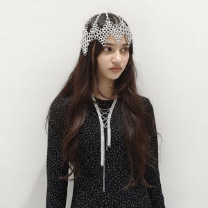 以下が含まれることがあります： 幾何学模様のシルバーのヘッドピース。小さな金属片が繋がって作られています。複数のチェーンが付いた長いシルバーのネックレスも見える。白い水玉模様の黒い長袖シャツを着ています。