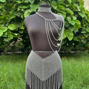 Puede incluir: Un conjunto de cota de malla plateada exhibido en un maniqu&iacute;. El conjunto incluye un collar&iacute;n, una pieza para el hombro con cadenas colgantes y una falda con dobladillo con flecos. El maniqu&iacute; est&aacute; sobre un fondo de follaje y hierba verdes.