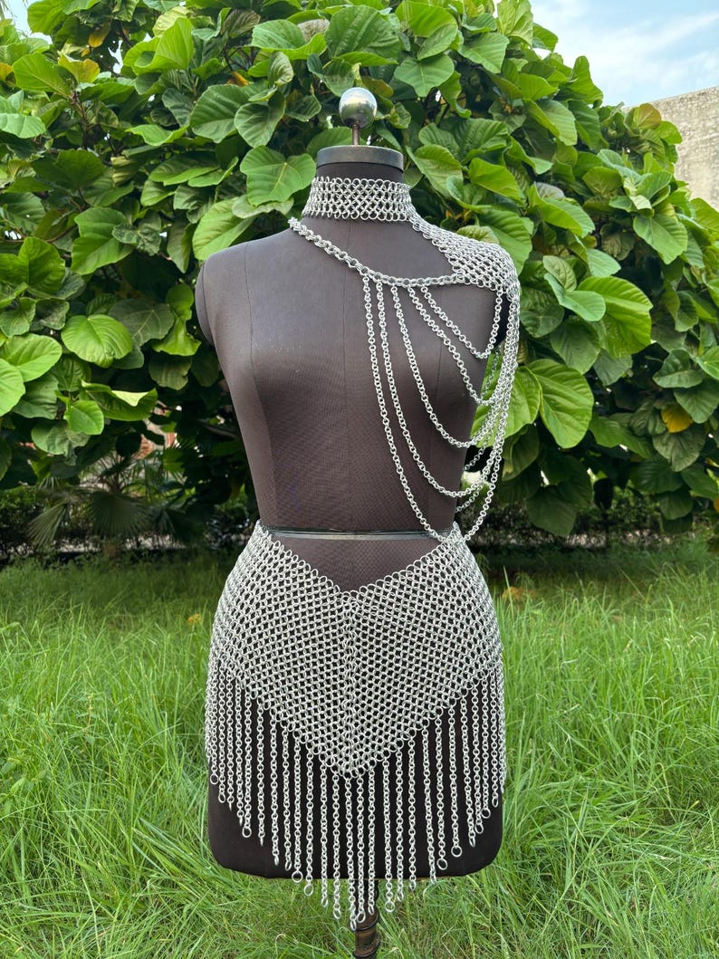 Puede incluir: Un conjunto de cota de malla plateada exhibido en un maniqu&iacute;. El conjunto incluye un collar&iacute;n, una pieza para el hombro con cadenas colgantes y una falda con dobladillo con flecos. El maniqu&iacute; est&aacute; sobre un fondo de follaje y hierba verdes.