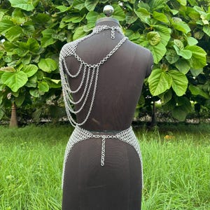 Puede incluir: Conjunto de top y falda de cota de malla plateada, exhibido en un maniqu&iacute; negro. El top presenta un cuello halter y cadenas en cascada sobre el hombro. La falda es un dise&ntilde;o ajustado a la cadera con un detalle de cadena. El conjunto est&aacute; hecho de peque&ntilde;os anillos met&aacute;licos interconectados.