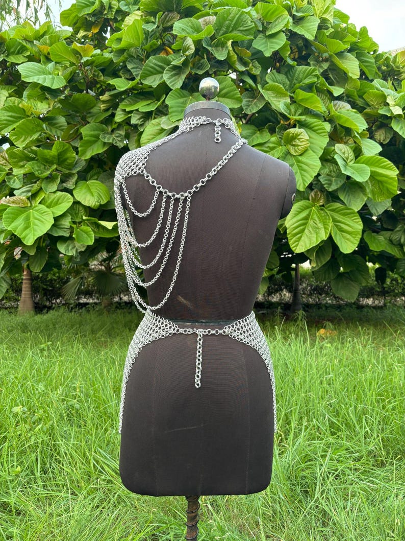 Puede incluir: Conjunto de top y falda de cota de malla plateada, exhibido en un maniqu&iacute; negro. El top presenta un cuello halter y cadenas en cascada sobre el hombro. La falda es un dise&ntilde;o ajustado a la cadera con un detalle de cadena. El conjunto est&aacute; hecho de peque&ntilde;os anillos met&aacute;licos interconectados.