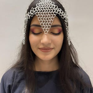 Puede incluir: Una diadema de cota de malla plateada con un dise&ntilde;o geom&eacute;trico. La diadema est&aacute; hecha de peque&ntilde;os anillos de metal entrelazados y se usa en la frente. La diadema tiene detalles de cadena colgantes en los lados.