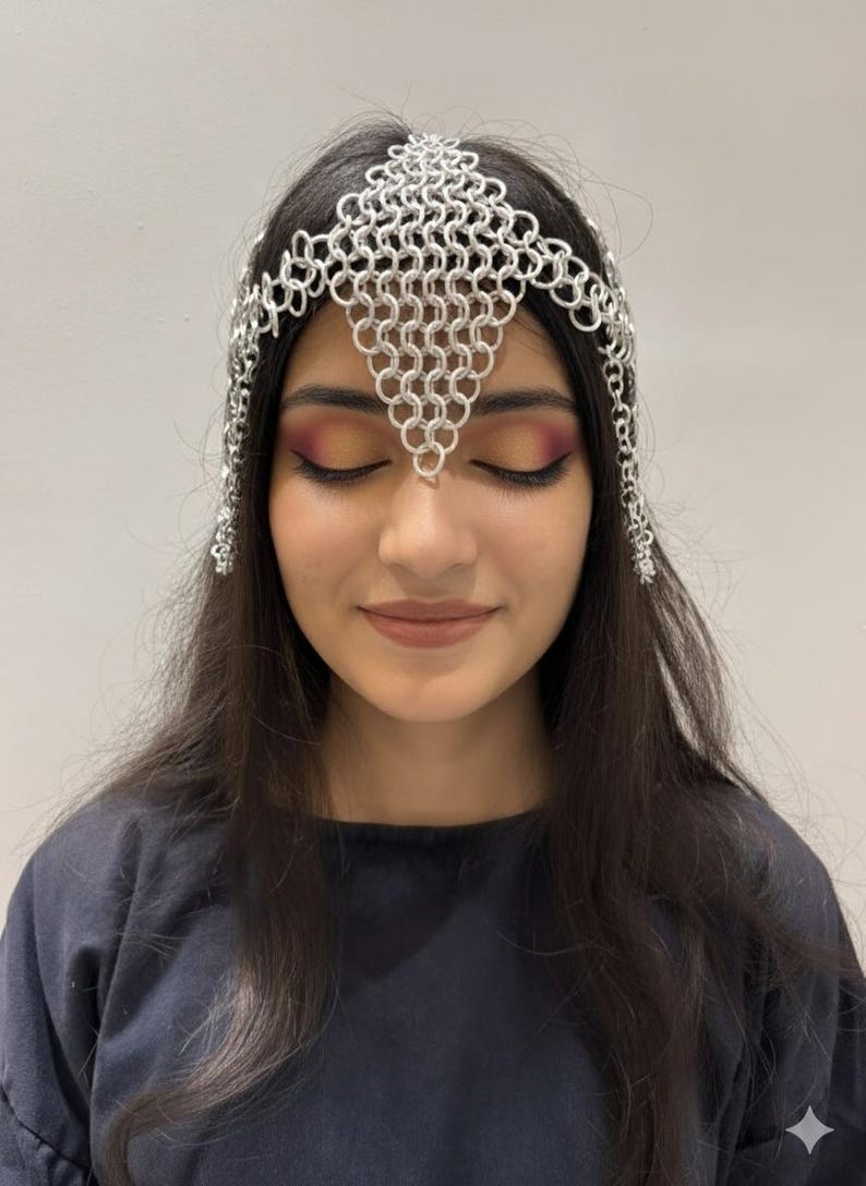 Puede incluir: Una diadema de cota de malla plateada con un dise&ntilde;o geom&eacute;trico. La diadema est&aacute; hecha de peque&ntilde;os anillos de metal entrelazados y se usa en la frente. La diadema tiene detalles de cadena colgantes en los lados.