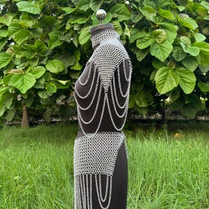 Puede incluir: Un conjunto de top y falda de cota de malla plateada exhibido en un maniqu&iacute; negro. La cota de malla est&aacute; hecha de peque&ntilde;os anillos de metal entrelazados. El top tiene un cuello alto y drapeados en los hombros. La falda tiene un dise&ntilde;o asim&eacute;trico con cadenas colgantes. El fondo es verde.