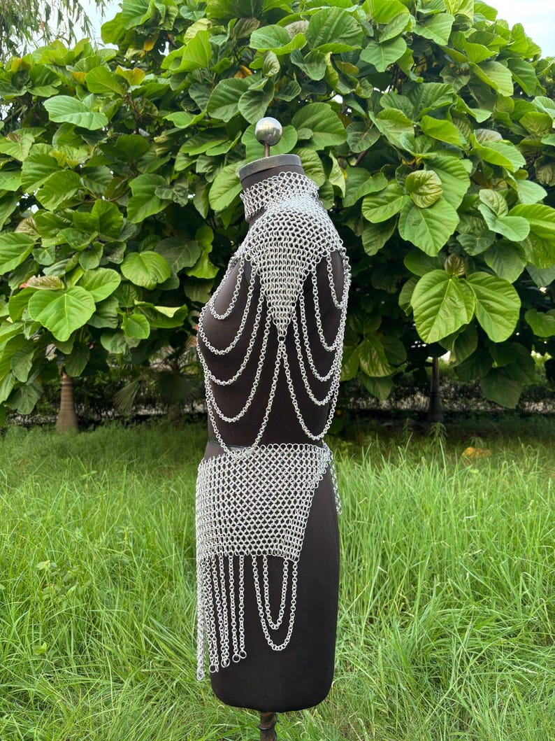 Puede incluir: Un conjunto de top y falda de cota de malla plateada exhibido en un maniqu&iacute; negro. La cota de malla est&aacute; hecha de peque&ntilde;os anillos de metal entrelazados. El top tiene un cuello alto y drapeados en los hombros. La falda tiene un dise&ntilde;o asim&eacute;trico con cadenas colgantes. El fondo es verde.